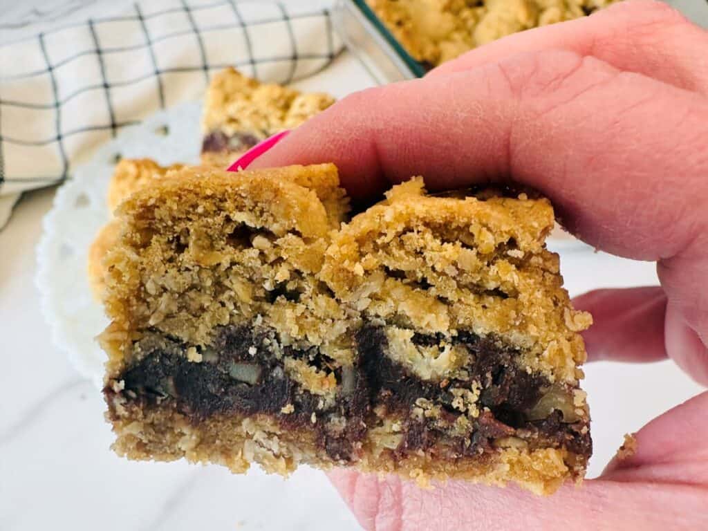 A hand holds a thick oat bar with a visible chocolate and nut filling, showing its crumbly texture. Other bars and a checkered towel are in the background.
