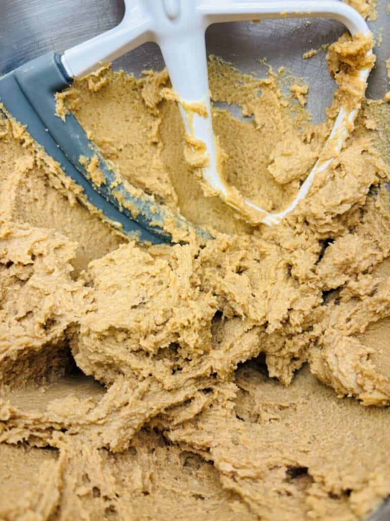 A close-up of cookie dough being mixed in a metal bowl with a stand mixer paddle attachment.