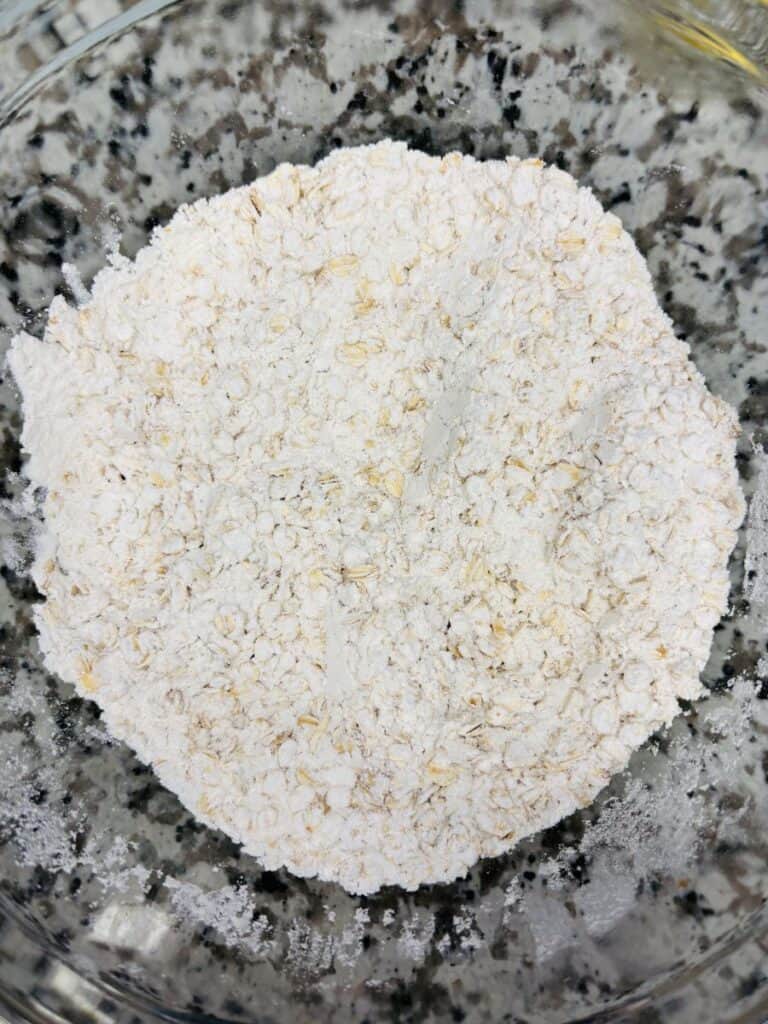 A glass bowl filled with a mixture of flour and oats sits on a speckled countertop, seen from above.
