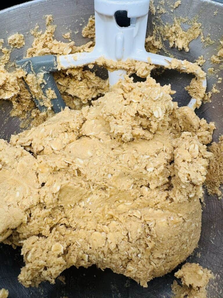Close-up of thick, tan-colored cookie dough with oats in a metal mixing bowl. A paddle attachment from an electric mixer is partially covered by the dough.