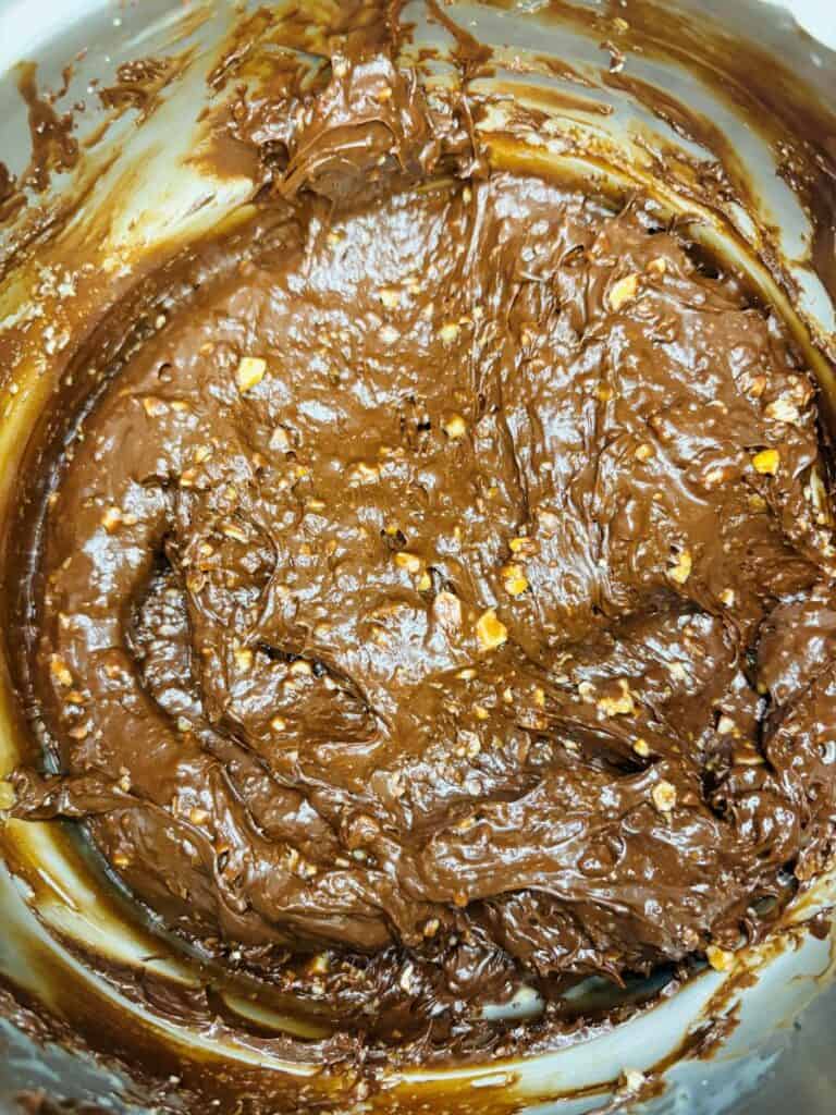 Close-up of thick, chocolate brownie batter with visible chunks of nuts in a mixing bowl. The batter has a glossy texture and is partially mixed.
