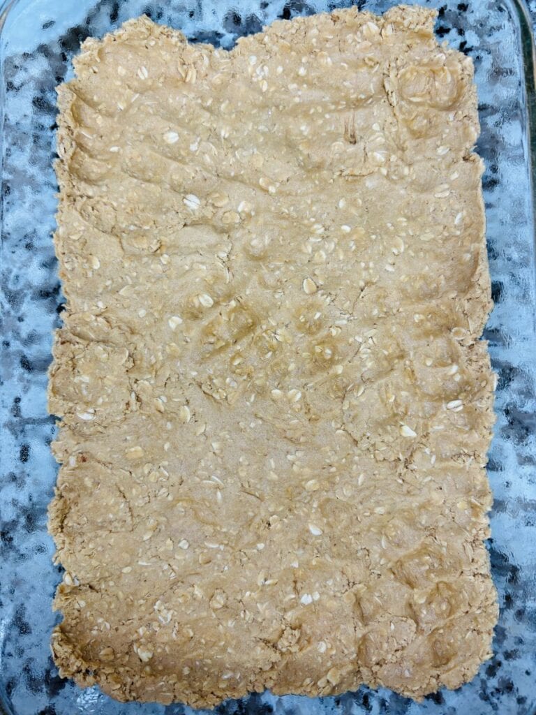 A rectangular glass baking dish filled with an unbaked, pressed oat mixture, likely for making bars or crust, with a textured surface.