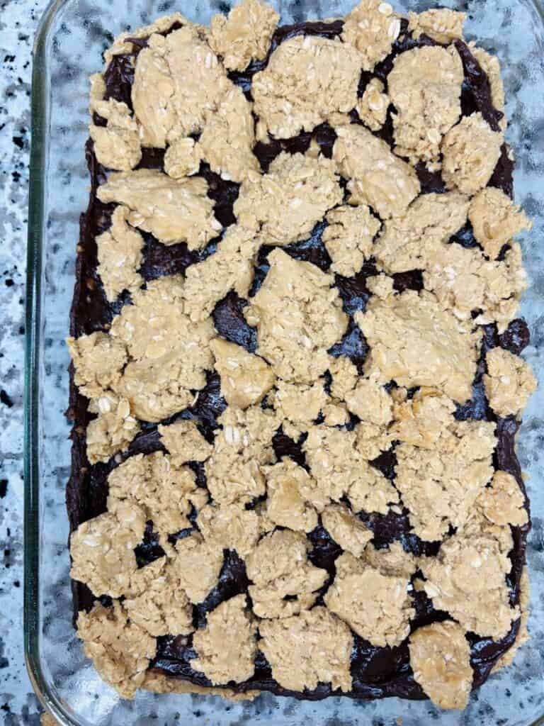 A glass baking dish with a dessert featuring a dark chocolate layer topped with uneven, crumbly oat and peanut butter mixture, sitting on a speckled granite countertop.