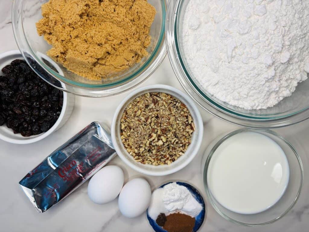Baking ingredients on a counter: brown sugar, flour, raisins, chopped nuts, butter, two eggs, milk, baking soda, cinnamon, and salt in bowls and containers.