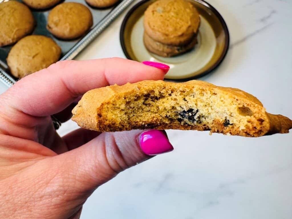 A hand with bright pink nails holds a half-eaten chocolate chip cookie. A plate and tray with more cookies are in the background on a white surface.