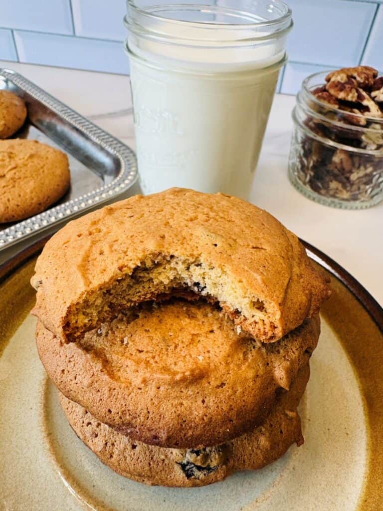 A stack of large cookies, with a bite taken out of the top one, sits on a plate. Behind them are a glass of milk, a small jar of pecans, and a tray with more cookies.
