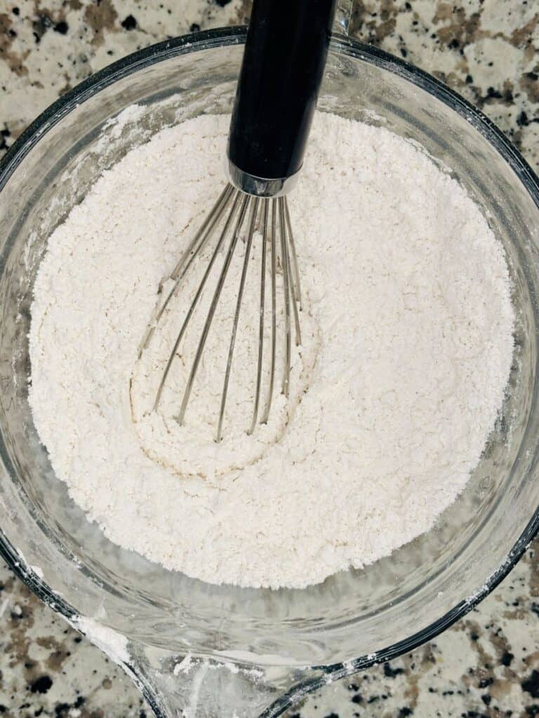 A metal whisk is mixing flour in a clear glass bowl on a speckled countertop.