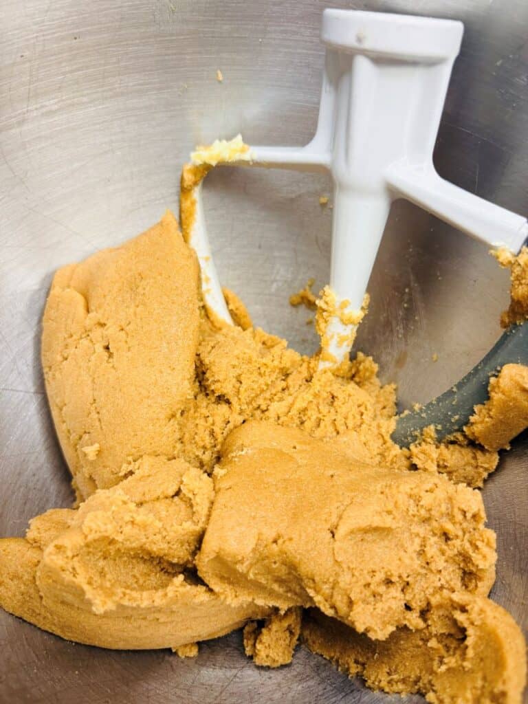 Cookie dough is being mixed in a metal bowl with a white paddle attachment on a stand mixer. The dough is light brown and appears thick and smooth.