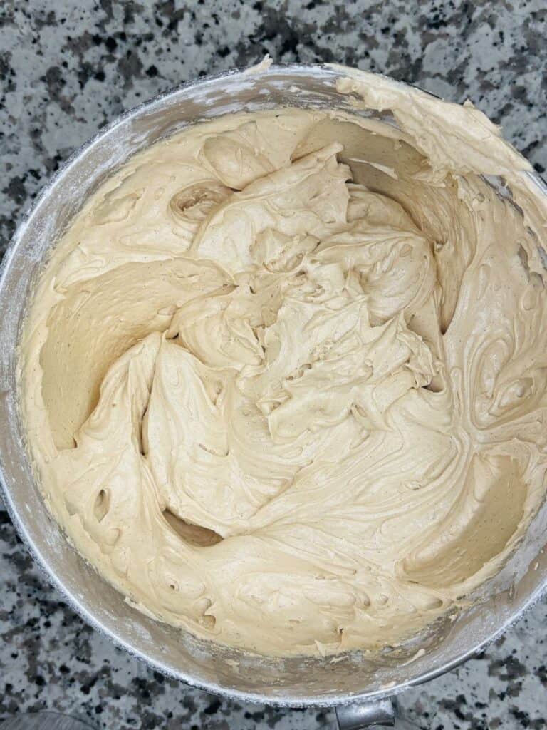 Top view of a mixing bowl filled with creamy, light brown batter or frosting, set on a speckled granite countertop.