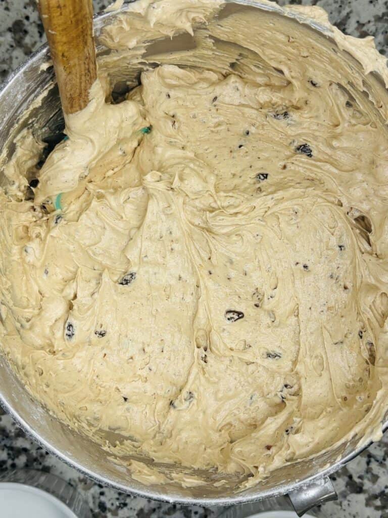 A metal mixing bowl filled with thick, light brown cookie dough with visible chocolate chips or raisins. A wooden spoon is partially submerged in the dough. The bowl sits on a speckled countertop.