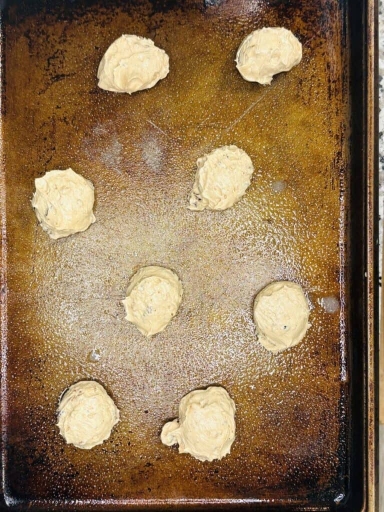 Eight scoops of cookie dough are spaced on a greased, well-used baking sheet, ready to be baked. The dough appears light brown with some darker flecks, likely chocolate chips or nuts.
