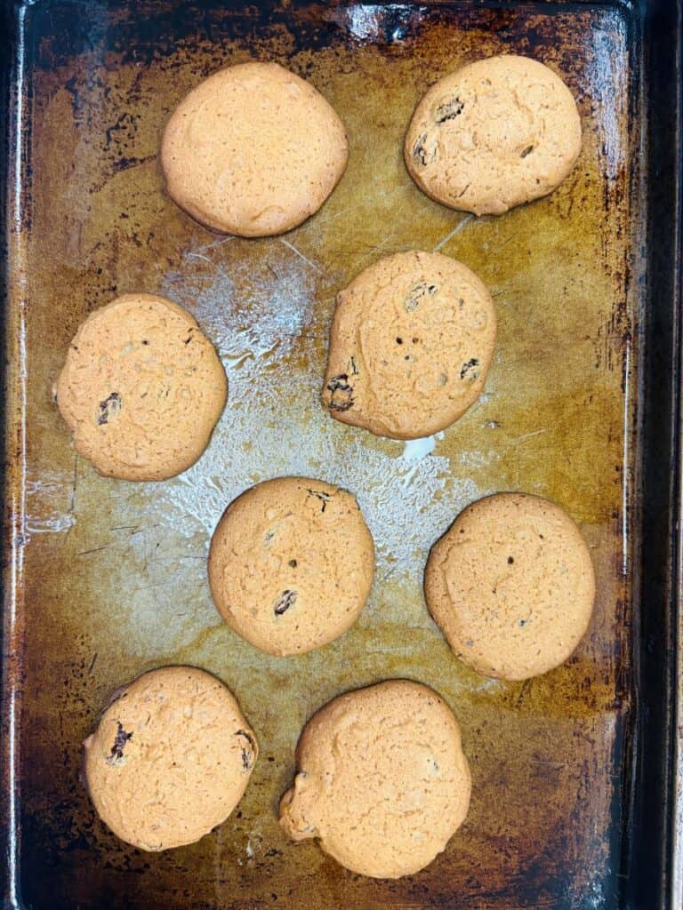 Eight golden brown cookies with some dark spots, likely raisins or chocolate chips, are spread out on a well-used, slightly scratched baking sheet.