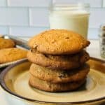 Four cookies stacked on a plate in front of a glass of milk, a jar of pecans, and a tray with more cookies. White tile wall in the background.