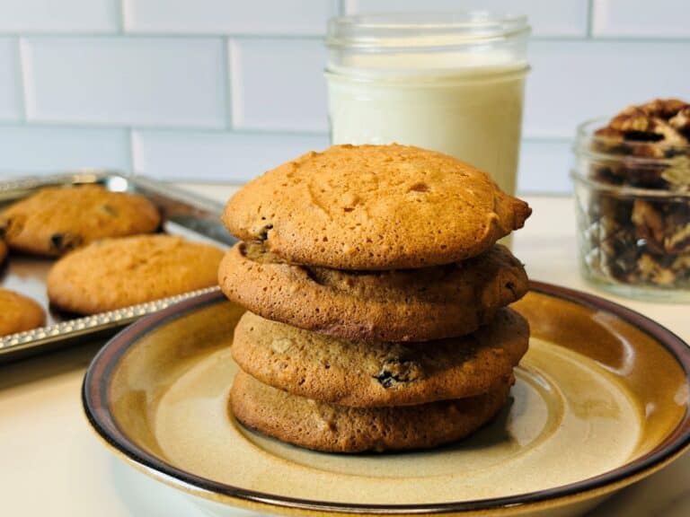 Four cookies stacked on a plate in front of a glass of milk, a jar of pecans, and a tray with more cookies. White tile wall in the background.