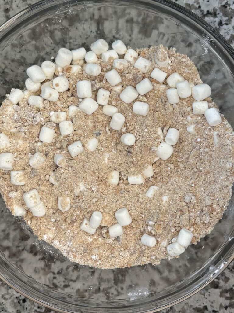 A glass bowl filled with a mixture of dry oats, crumbs, and small white mini marshmallows sits on a gray granite countertop.