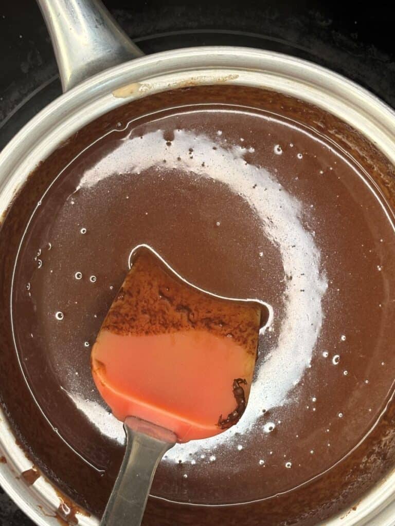 A red spatula stirs glossy melted chocolate in a metal saucepan, with swirls and bubbles visible on the surface.