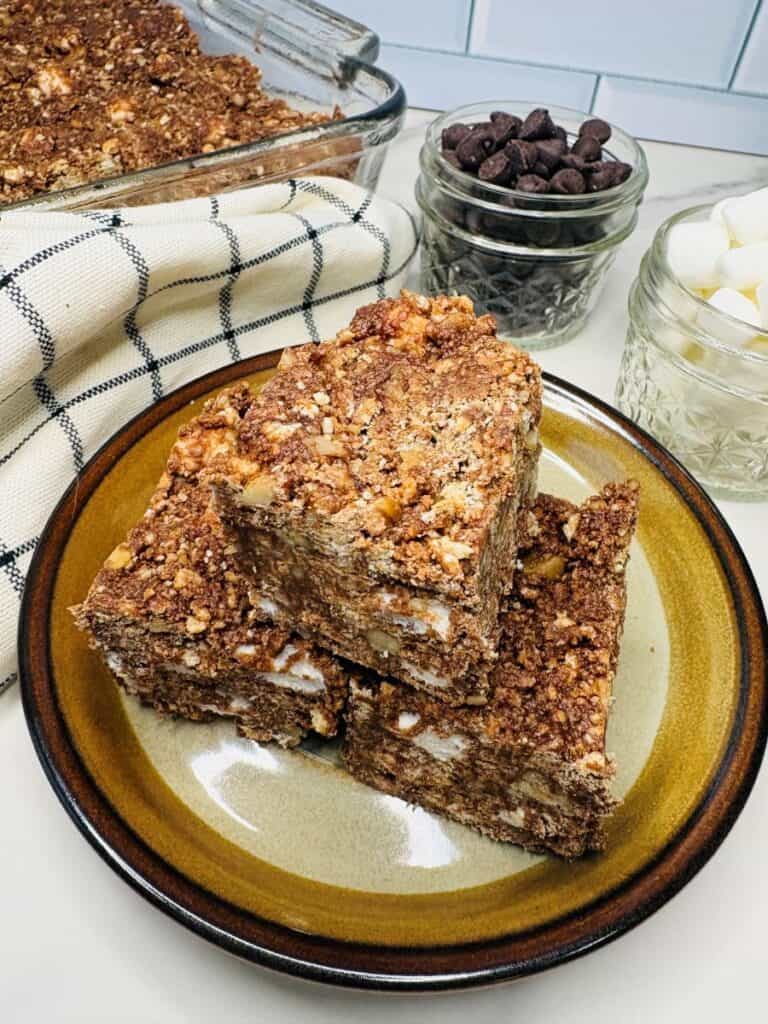 Three chocolate and marshmallow dessert bars are stacked on a plate, with a baking dish, a jar of chocolate chips, marshmallows, and a checkered towel in the background.