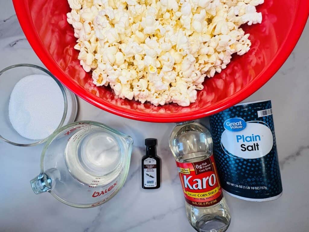 Large red bowl of popcorn with ingredients: sugar, vanilla extract, corn syrup, plain salt, and a measuring cup of water on a marble surface.