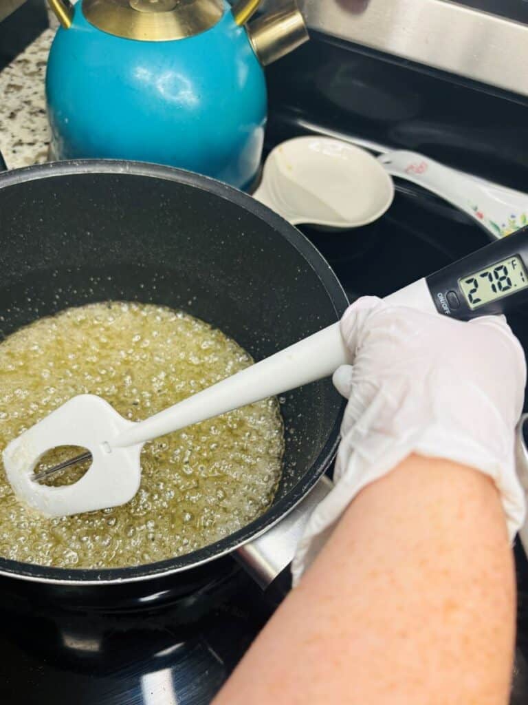 A gloved hand holds a spatula and a candy thermometer in a bubbling pot of sugar syrup on a stovetop. The thermometer reads 218°F. A blue kettle and white bowls are in the background.