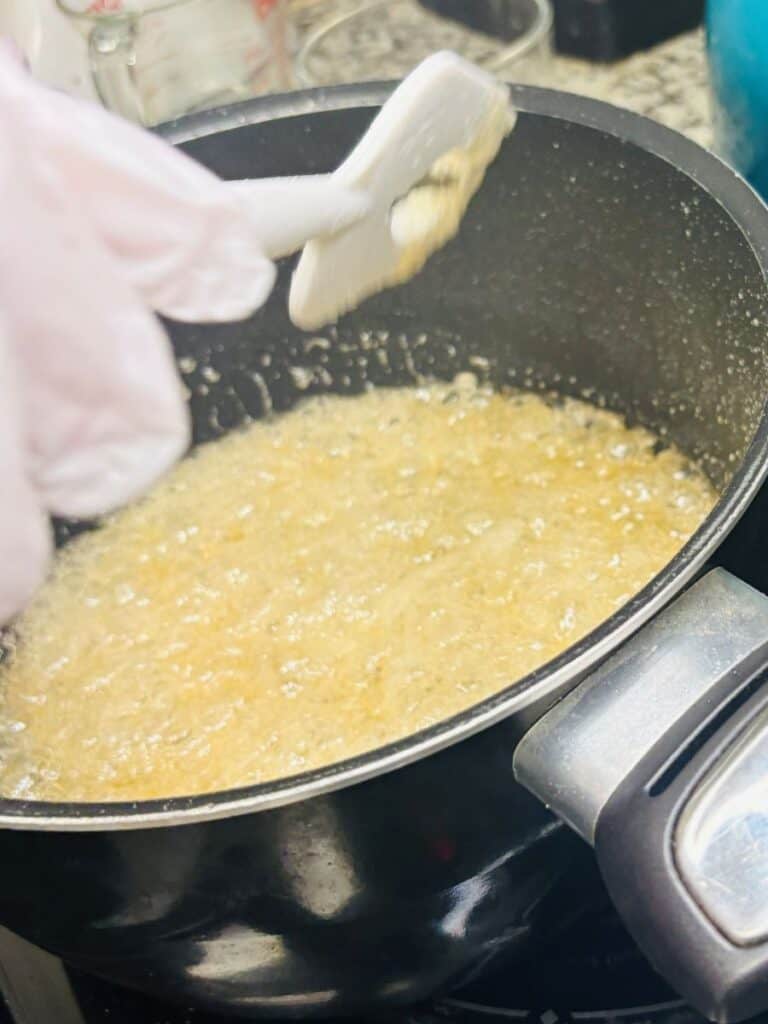 A pot of bubbling caramel is being stirred with a white spatula by a person wearing a white glove. The scene is on a stovetop with kitchen tools visible in the background.