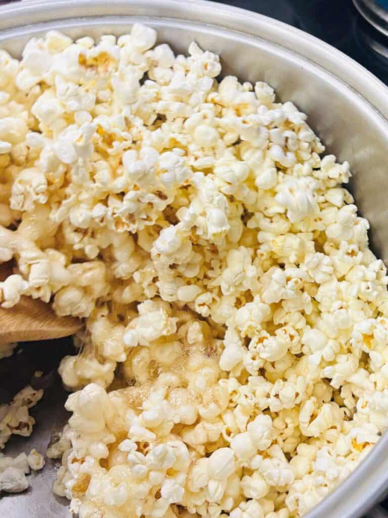 A close-up of freshly popped popcorn in a metal pot, with melted butter pooling at the bottom and a wooden spoon partially visible on the left side.