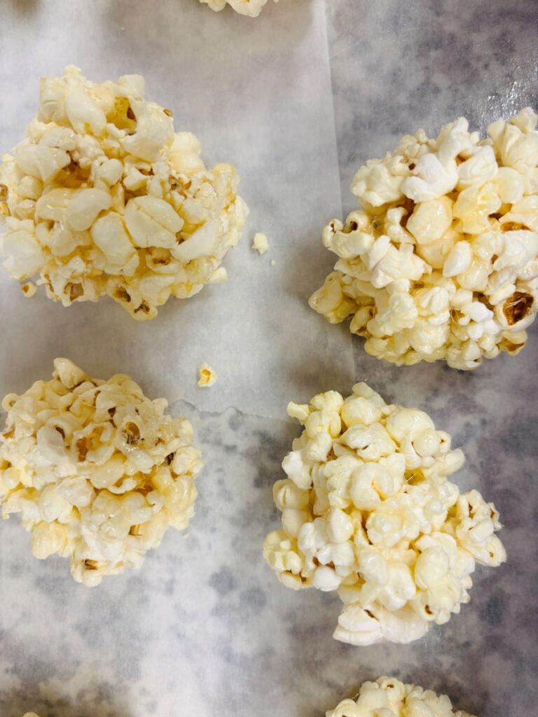 Four popcorn balls placed on parchment paper, each made from popped corn kernels stuck together, viewed from above. The surface beneath them is lightly speckled.