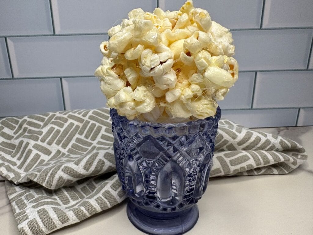 A purple glass cup filled to the top with popped popcorn sits on a countertop in front of a gray patterned towel and white subway tile backsplash.