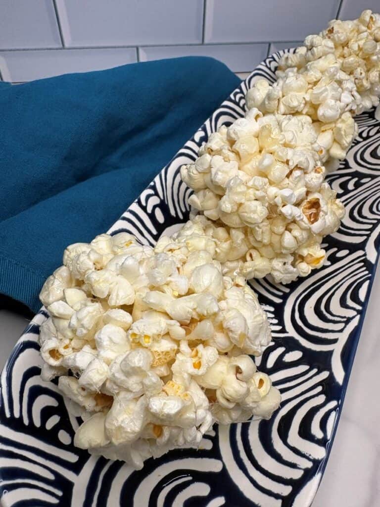 Three popcorn balls sit on a long, patterned black-and-white platter next to a folded dark teal napkin, all set against a white tile background.