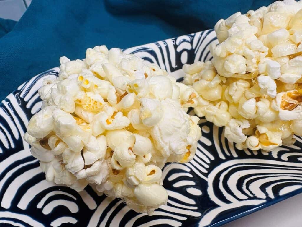 Two popcorn balls rest on a patterned black and white plate, with a blue cloth in the background.