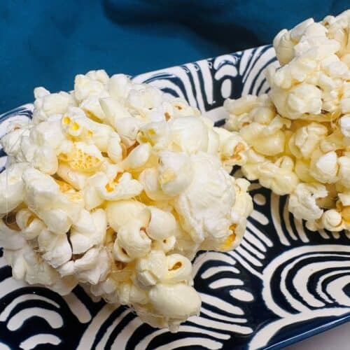 Two popcorn balls rest on a patterned black and white plate, with a blue cloth in the background.