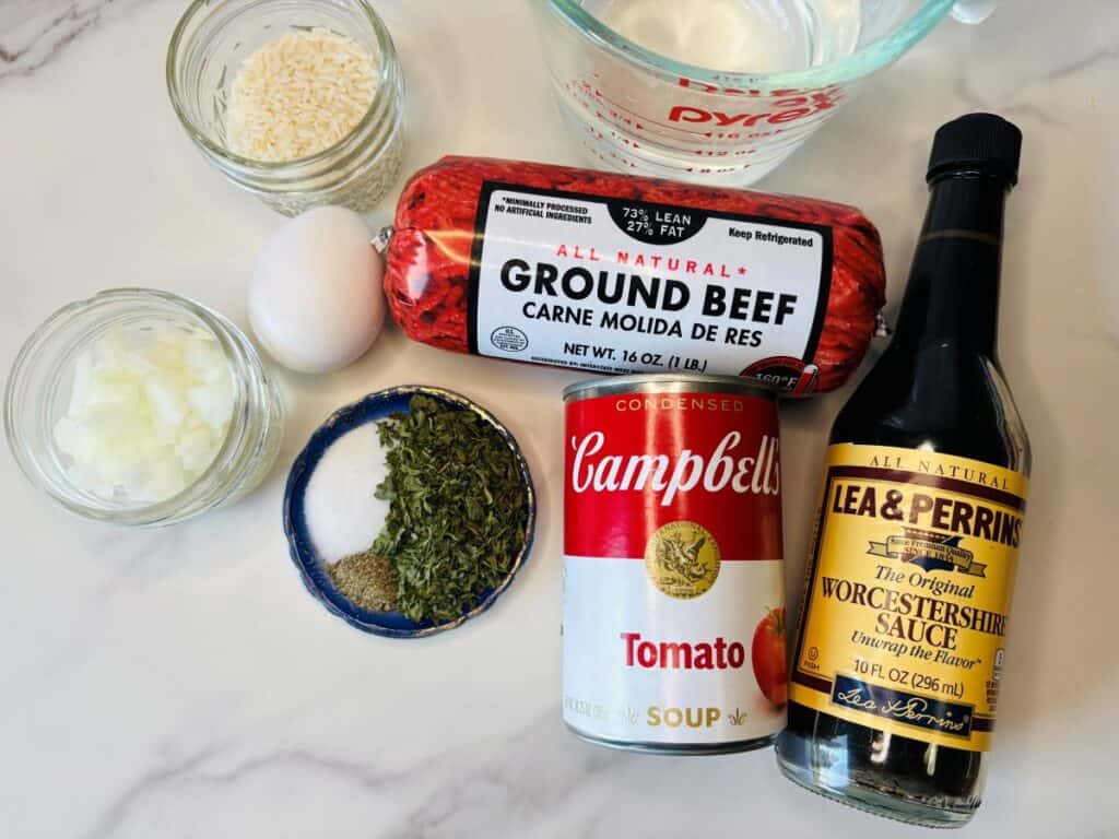 Ingredients on a marble counter: ground beef, tomato soup can, Worcestershire sauce, egg, chopped onion, white rice, water, and a dish with salt and dried herbs.