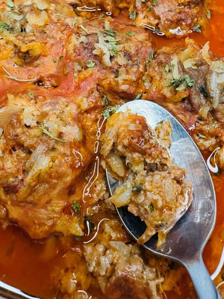 A close-up of a spoon lifting a portion of meatloaf in tomato sauce, showing chunks of meat, herbs, and onions in a rich, red, oily liquid.