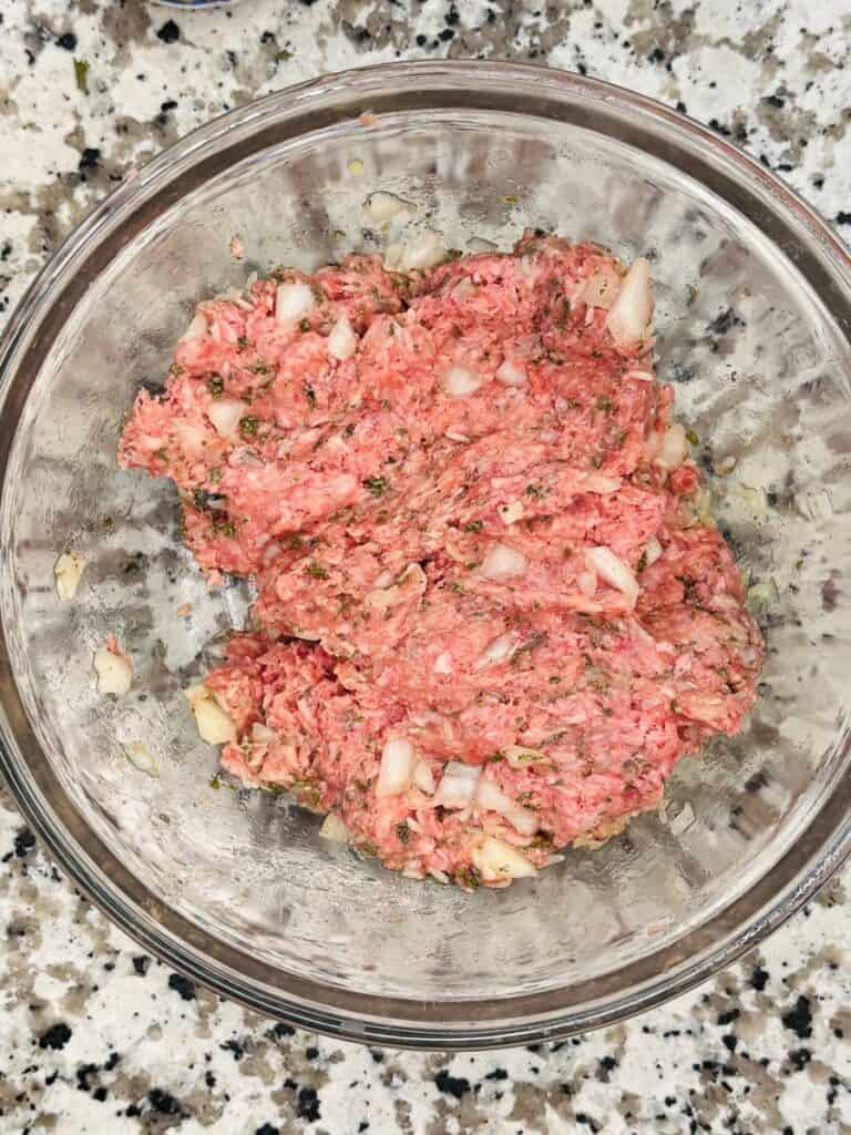 A glass bowl filled with raw ground meat, chopped onions, and herbs, sitting on a granite countertop.