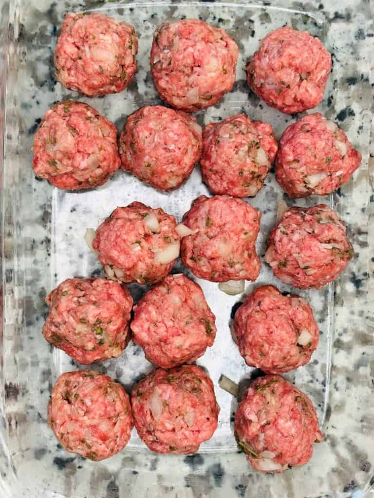 Raw meatballs arranged in a clear glass baking dish, ready to be cooked. The meat mixture includes visible herbs and small onion pieces.