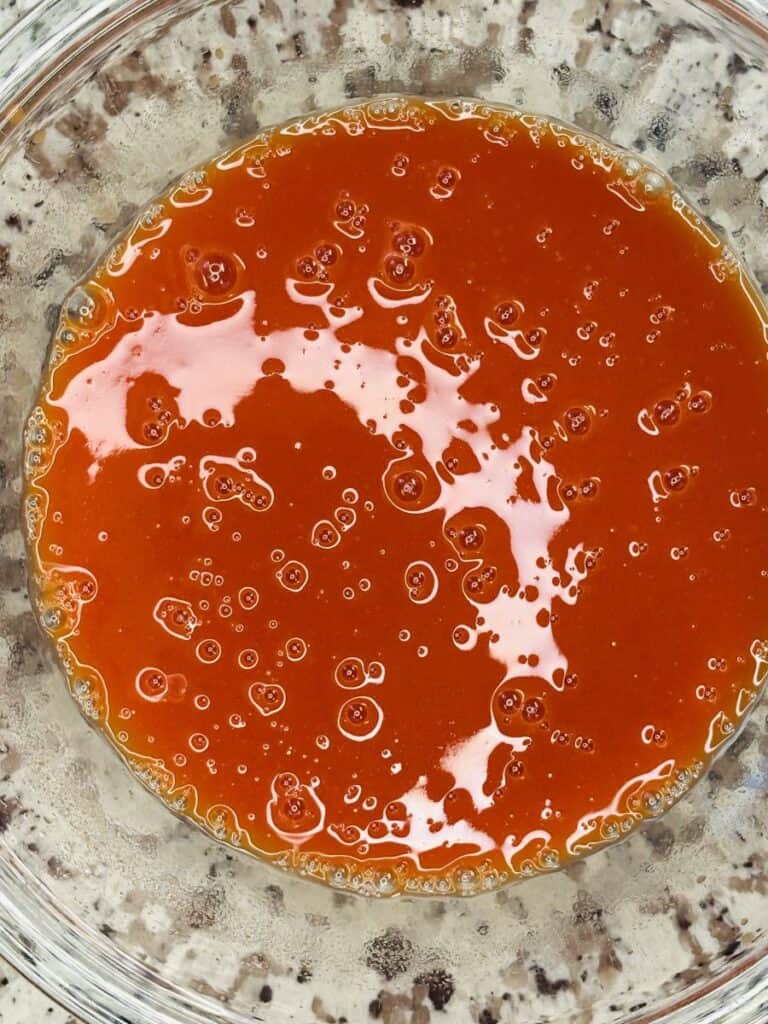 A clear glass bowl filled with a glossy, orange-red liquid with small bubbles on the surface, sitting on a speckled countertop.