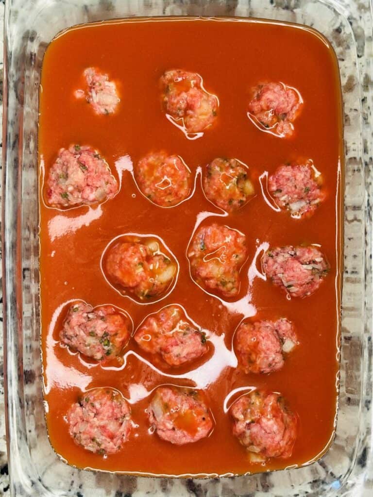 Uncooked meatballs in tomato sauce arranged in a clear glass baking dish, ready to be baked. The sauce covers the bottom, and the meatballs are spaced evenly throughout the dish.