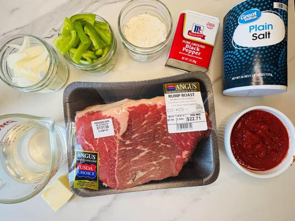 Raw rump roast, green bell peppers, onion slices, flour, ground black pepper, plain salt, tomato sauce, a measuring cup of water, and a cube of butter arranged on a white countertop.