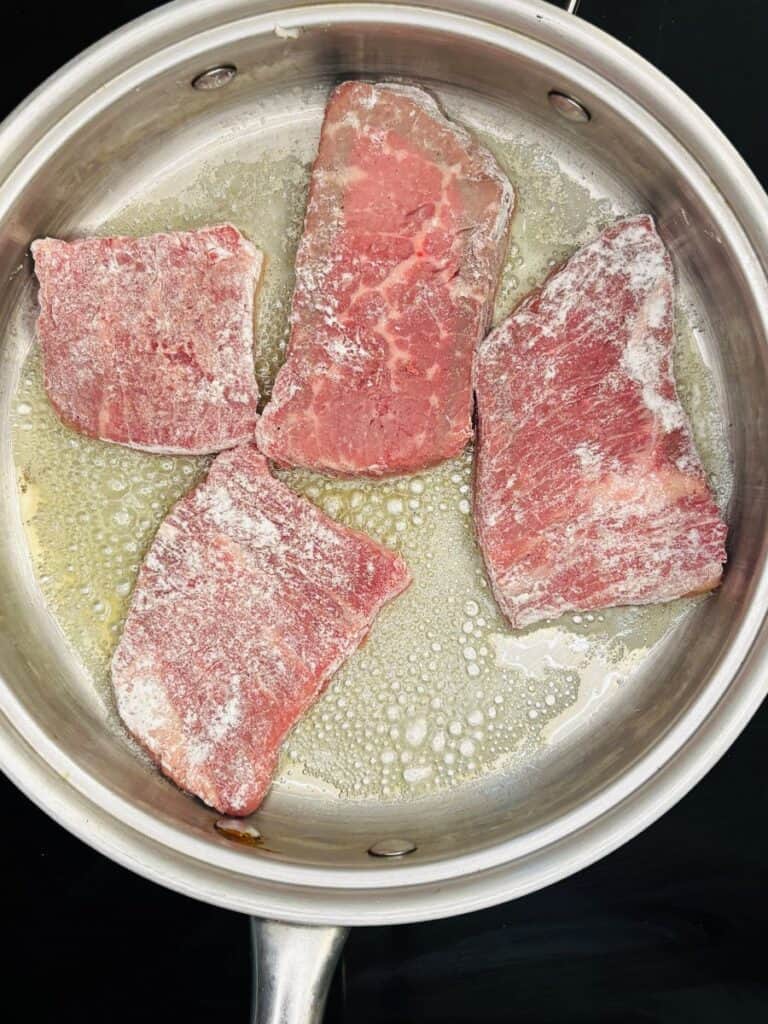 Four raw, flour-coated beef pieces sizzling in melted butter inside a stainless steel pan on a stovetop.