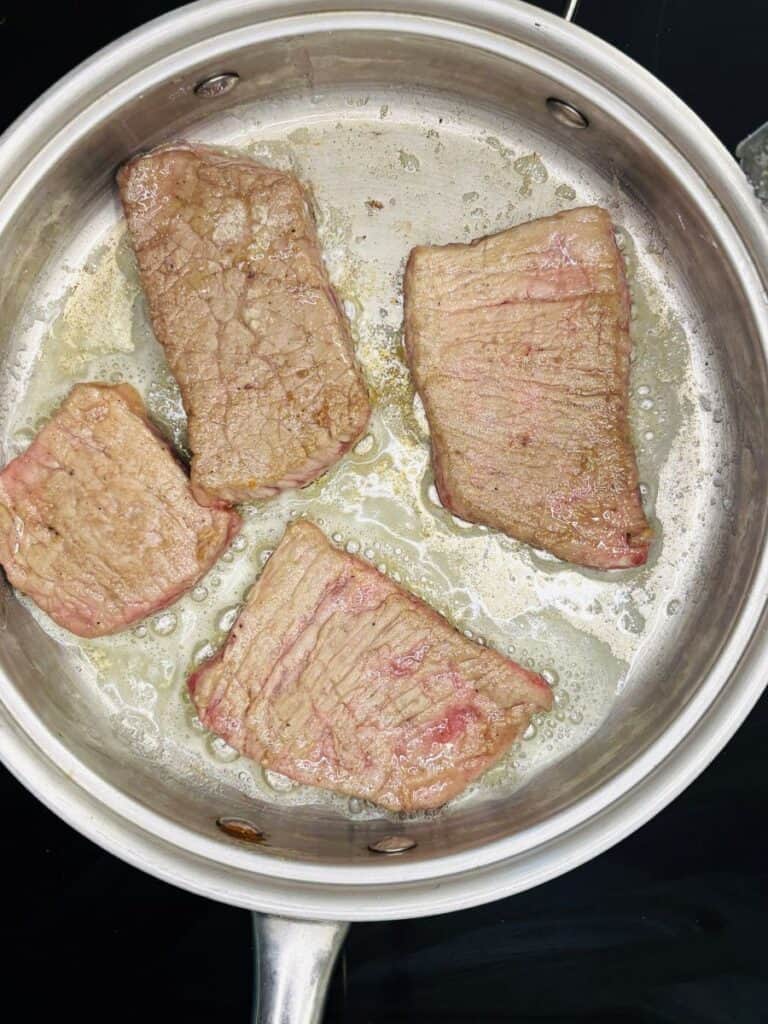 Four pieces of beef are searing in a stainless steel pan with bubbling oil. The meat is browned on the outside, with some pink areas still visible, indicating it is partially cooked.