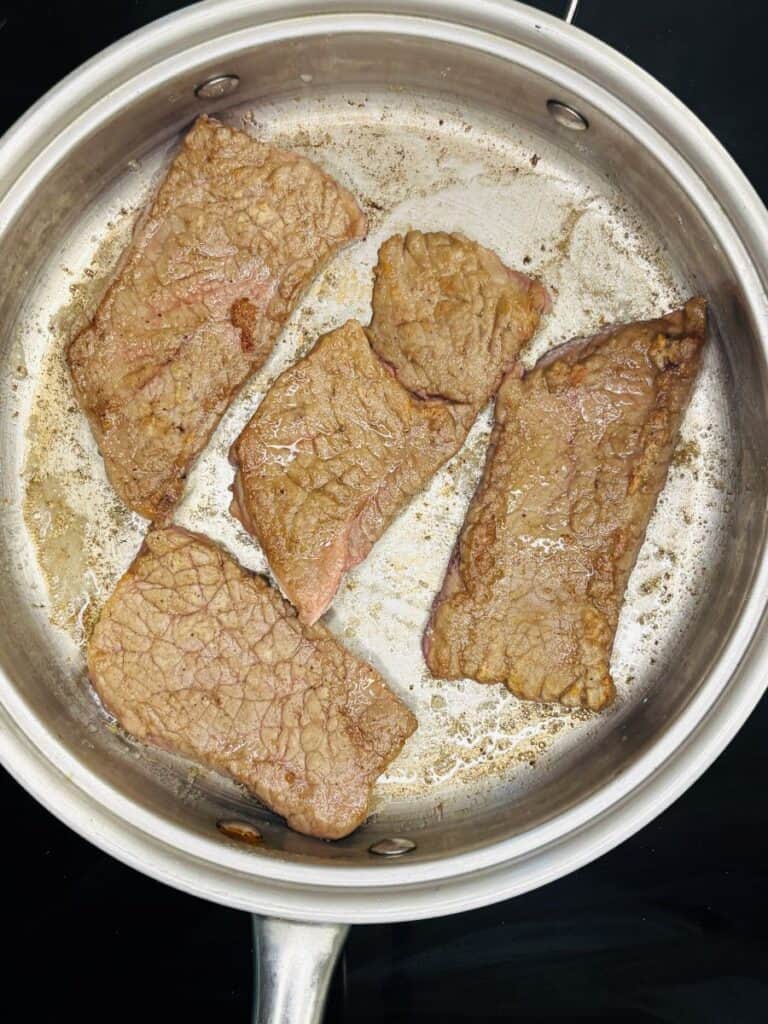 Four browned beef steak strips cooking in a stainless steel frying pan. The pan is on a stove, and the meat appears seared with some juices and browning on the pan surface.
