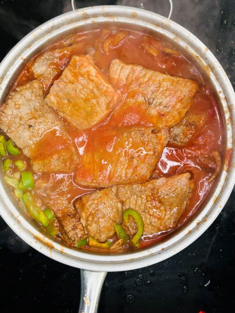 Slices of beef simmer in a red tomato sauce with green bell pepper rings in a silver pot on a stovetop.