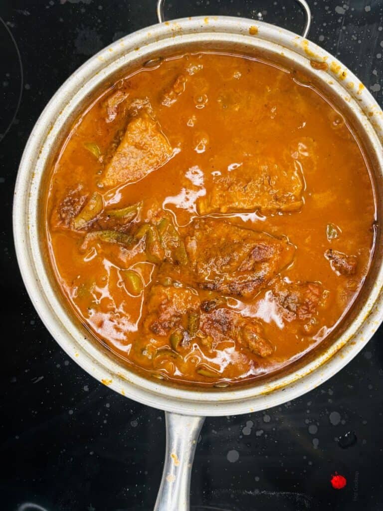 A stainless steel pot filled with rich, reddish-brown stew containing chunks of meat and green peppers, simmering on a black stovetop.