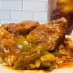 A plate of rice topped with pork chops, green peppers, and tomato-based sauce, with a glass of iced tea in the background.
