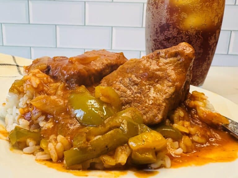 A plate of rice topped with pork chops, green peppers, and tomato-based sauce, with a glass of iced tea in the background.