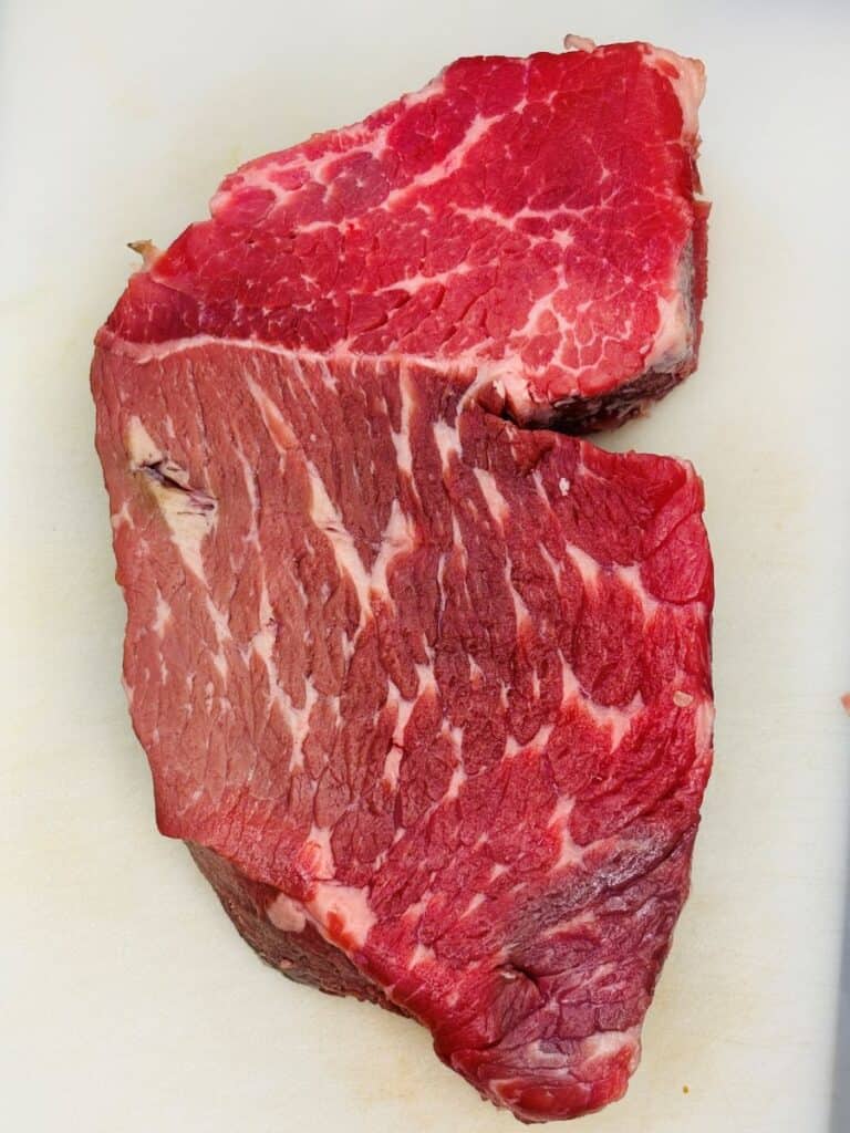 A raw, marbled piece of beef steak sits on a white cutting board. The meat shows visible veins of fat and varying shades of red.