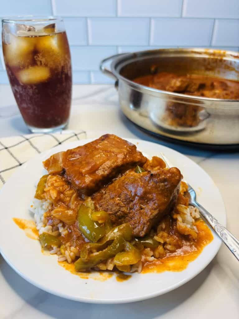 Plate of smothered steak with gravy and peppers over rice, next to a fork. A glass of iced drink and a pot with more steak are in the background on a white countertop with a white tile backsplash.