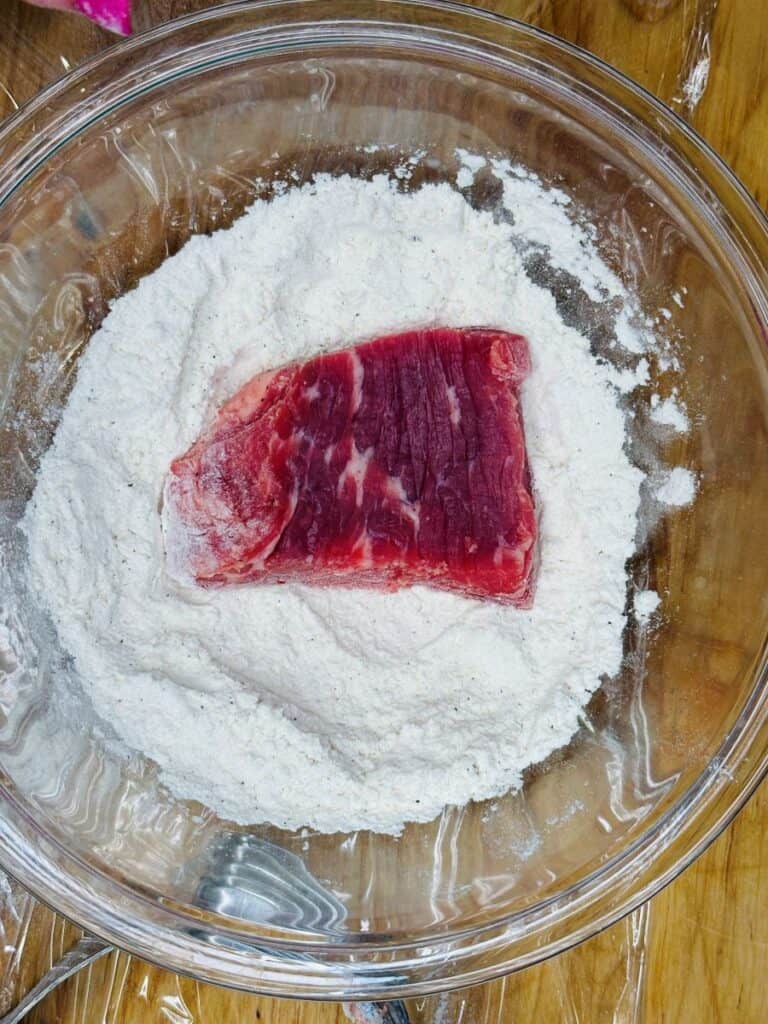A raw slice of beef sits on top of a mound of flour in a clear glass bowl, placed on a wooden surface.