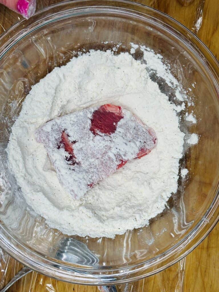 A raw steak coated with flour sits in a large clear glass bowl filled with flour, on a wooden surface. A metal fork is partially visible at the bottom of the bowl.