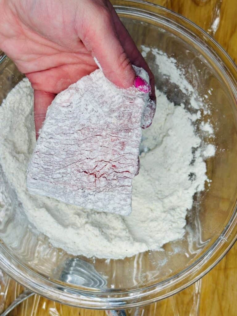 A hand with pink nail polish holds a piece of raw meat coated in flour over a glass bowl filled with flour on a wooden surface.