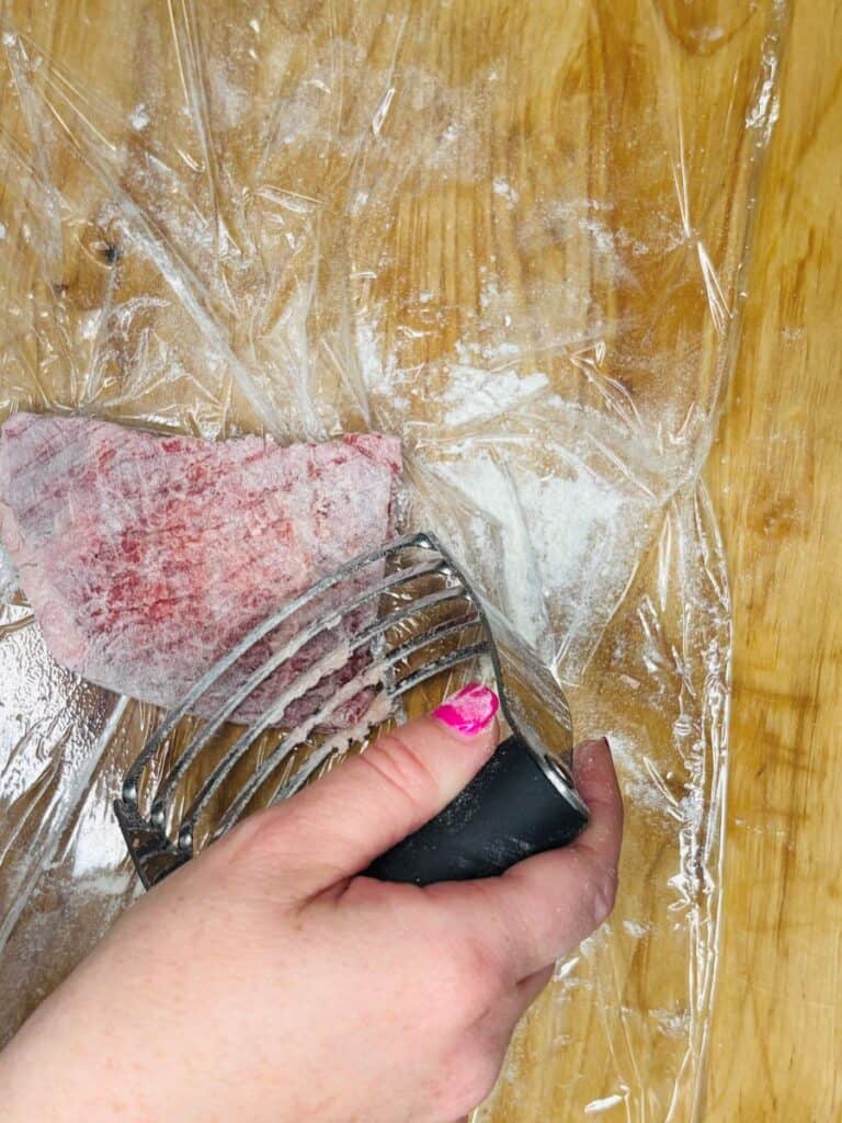 A hand with pink nail polish uses a meat tenderizer on a piece of raw meat covered in plastic wrap on a wooden surface.